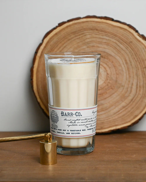 A Barr-Co. Original Scent Parfait Candle sits on a wooden surface with a tree slice in the background and a gold snuffer nearby, offering a long-lasting burn in the comforting Barr-Co. signature fragrance.