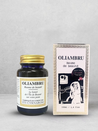 A jar and box of ISULA parfums Oliambru Beauty Balm, enriched with Corsican Immortelle and organic oils, rest on a light gray surface. The box shows a woman’s illustration; the jar has a gold lid with black and gold text.