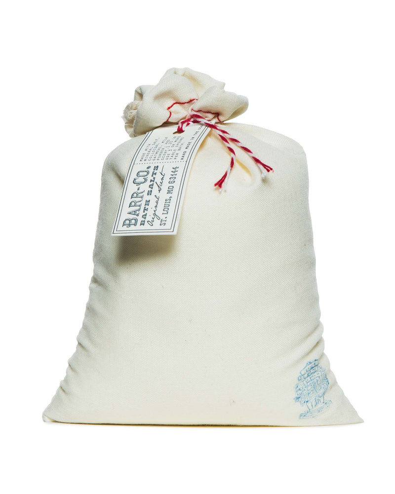 A cream-colored cloth bag, tied with red and white string, makes for a perfect relaxation gift. It contains the Barr-Co. Original Scent Bath Salt. The bag features a "BARR CO." tag with contact details and is adorned with a blue emblem stamped near its base.