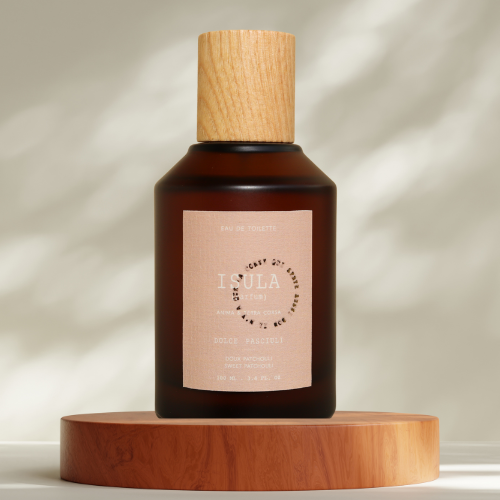 A dark amber glass perfume bottle with a light wooden cap sits on a round wooden platform. The label reads “ISULA parfums Dolce Pasciuli - Soft Patchouli eau de toilette,” against a neutral, softly lit background.