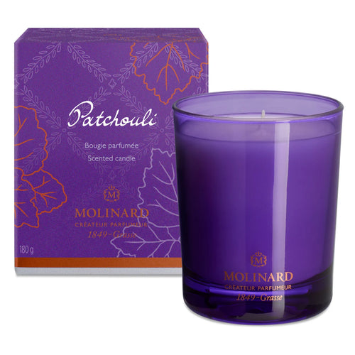 The Molinard Candle - Patchouly by Molinard is displayed in a purple glass holder before its matching purple and orange box, decorated with leaf outlines and product details, echoing the candle’s complex aroma.