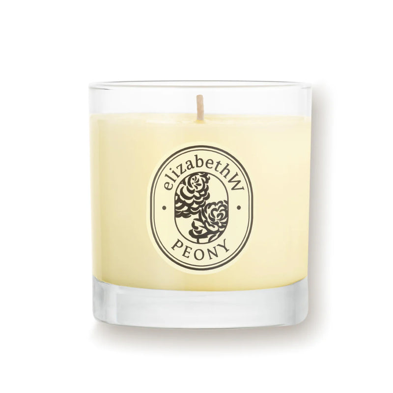 The "elizabeth W Botanist's Scent Lab Peony Candle" is presented in a clear glass holder adorned with an elegant floral design. The candle, made from renewable soy wax, features a light cream-colored wax with a centered wick that symbolizes purity against its plain white background.