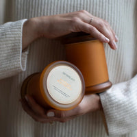 A person wearing a cream knit sweater holds two amber glass candles, including the limited edition Archipelago Botanicals Pumpkin Macchiato Jar Candle. A delicate ring adorns their hand.