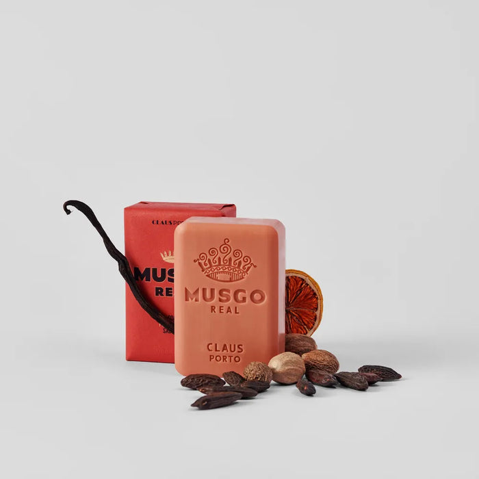 A bar of Claus Porto 1887 Musgo Real Puro Sangue Body Soap next to its packaging box, accompanied by a dried orange slice, vanilla pod, and assorted spices on a white background.