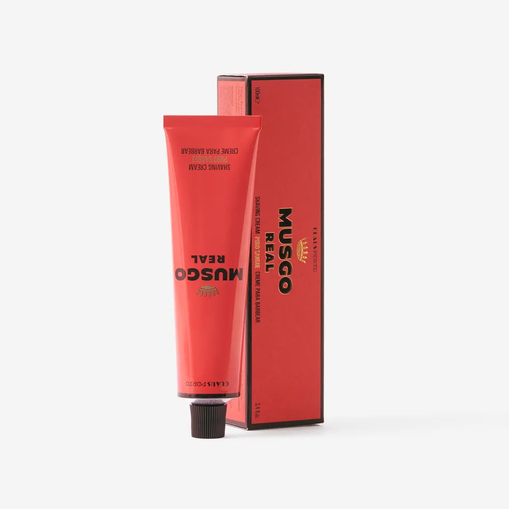 A red tube of Claus Porto Musgo Real Puro Sangue Shaving Cream by Claus Porto 1887 stands upright before its matching box, both labeled with product details. The lanolin cream offers a woody-leathery scent. The background is plain white.