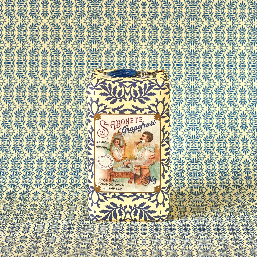 A bar of Claus Porto 1887 Classico Barbear Grapefruit Fig Soap, wrapped in blue and white patterned paper, stands upright against a coordinating background. The label shows vintage art of a woman by a mirror and reads "Sabonete Grapefruit Fig Scent.