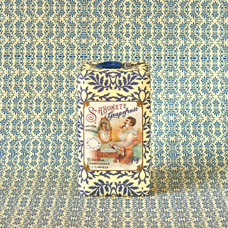 A bar of Claus Porto 1887 Classico Barbear Grapefruit Fig Soap, wrapped in blue and white patterned paper, stands upright against a coordinating background. The label shows vintage art of a woman by a mirror and reads "Sabonete Grapefruit Fig Scent.