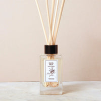 A Norfolk Natural Living Coastal Sea Salt Reed Diffuser, filled with oil and several wooden reeds, sits on a light surface against a beige background. The glass bottle features the Norfolk Natural Living label.
