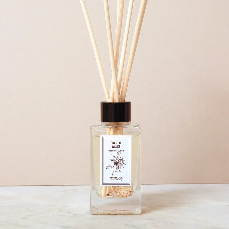 A Norfolk Natural Living Coastal Sea Salt Reed Diffuser, filled with oil and several wooden reeds, sits on a light surface against a beige background. The glass bottle features the Norfolk Natural Living label.