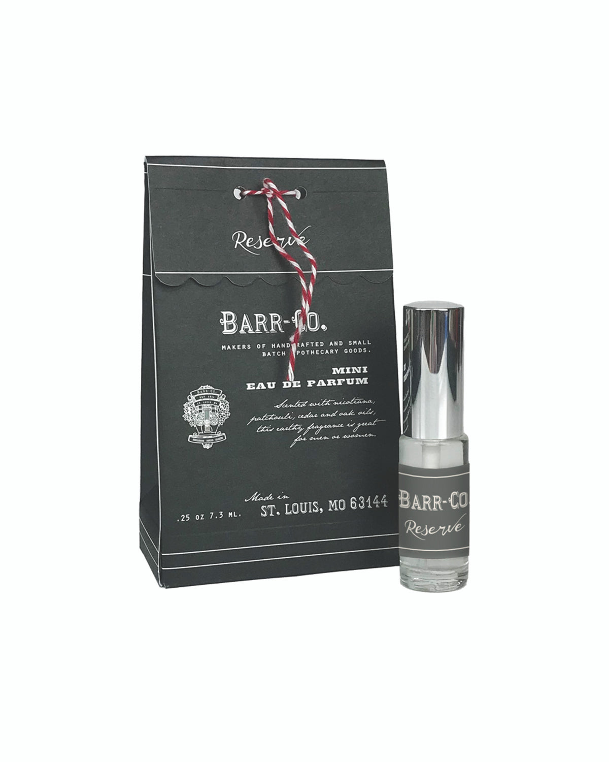 A bottle of Barr-Co. Reserve Mini-Perfume next to its black packaging bag, tied with a white and red twisted cord. The label includes brand and address details and the scent notes.