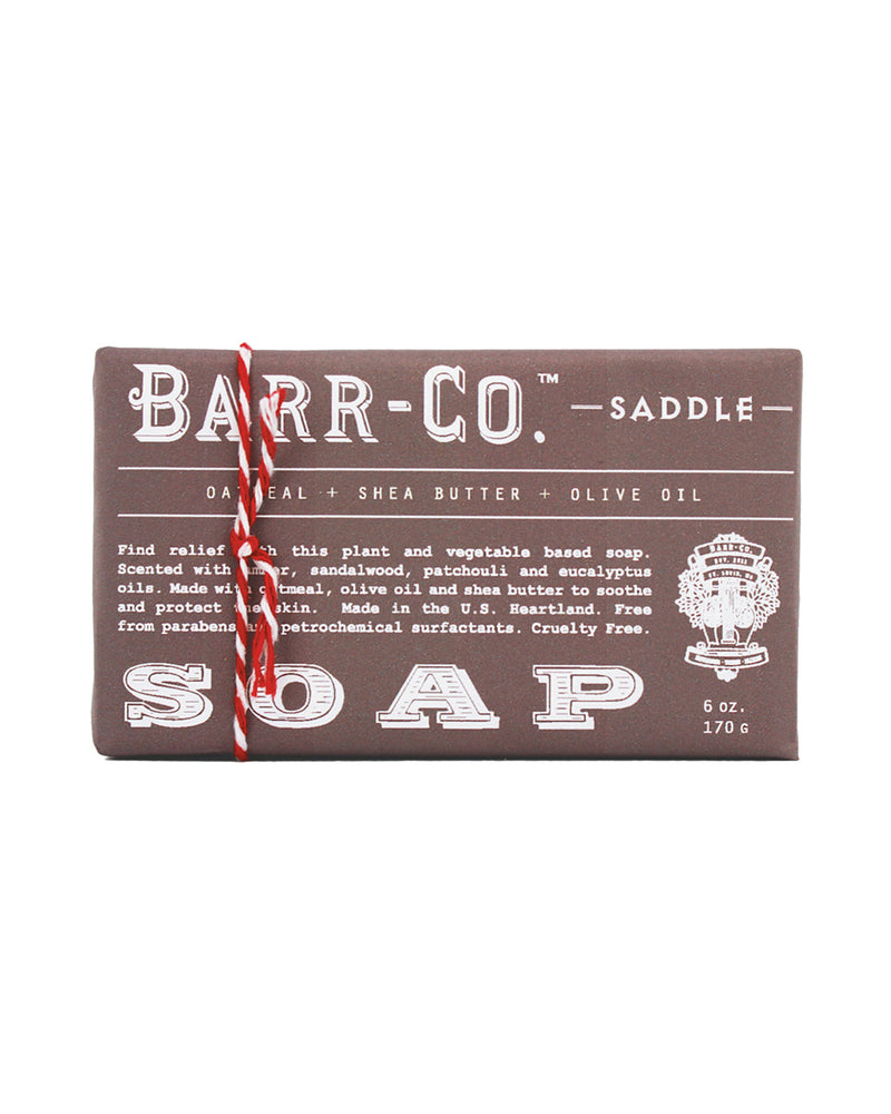 A bar of Barr-Co Saddle Triple-Milled Bar Soap wrapped in brown paper with white text. The packaging states the soap contains oatmeal, shea butter, and olive oil, and is scented with vetiver, sandalwood, patchouli, and eucalyptus essential oils. It is 6 oz (170 g) and cruelty free.