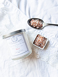 The Z&Co. Salt Farmhouse Candle, made with soy coconut wax, sits in a glass jar labeled "Salt" on a lace cloth next to a square dish and spoon filled with pink Himalayan salt crystals.