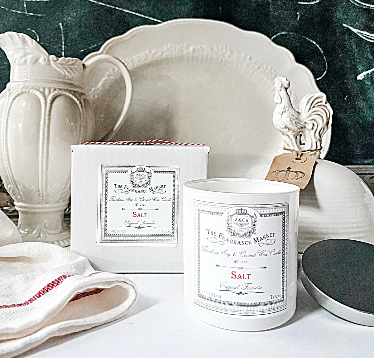 A Z&Co. Salt Farmhouse Candle made from soy coconut wax sits beside its box on a white countertop, surrounded by white ceramic dishes, a rooster figurine with a tag, and a white towel with red stripes.