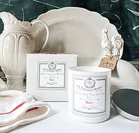 A Z&Co. Salt Farmhouse Candle made from soy coconut wax sits beside its box on a white countertop, surrounded by white ceramic dishes, a rooster figurine with a tag, and a white towel with red stripes.