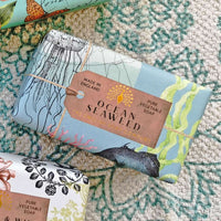An artisanal soap bar labeled "The English Soap Co. Anniversary Ocean Seaweed Soap" wrapped in aqua-colored paper with marine illustrations, bound by twine, displayed on a textured rug.