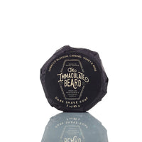 A round, black-wrapped "The Immaculate Beard DARK Shave Soap Puck" sits on a reflective surface. Scented with tobacco blossom, caramel, honey, and rose, it's perfect for sensitive skin and pairs well with your favorite shave brush.