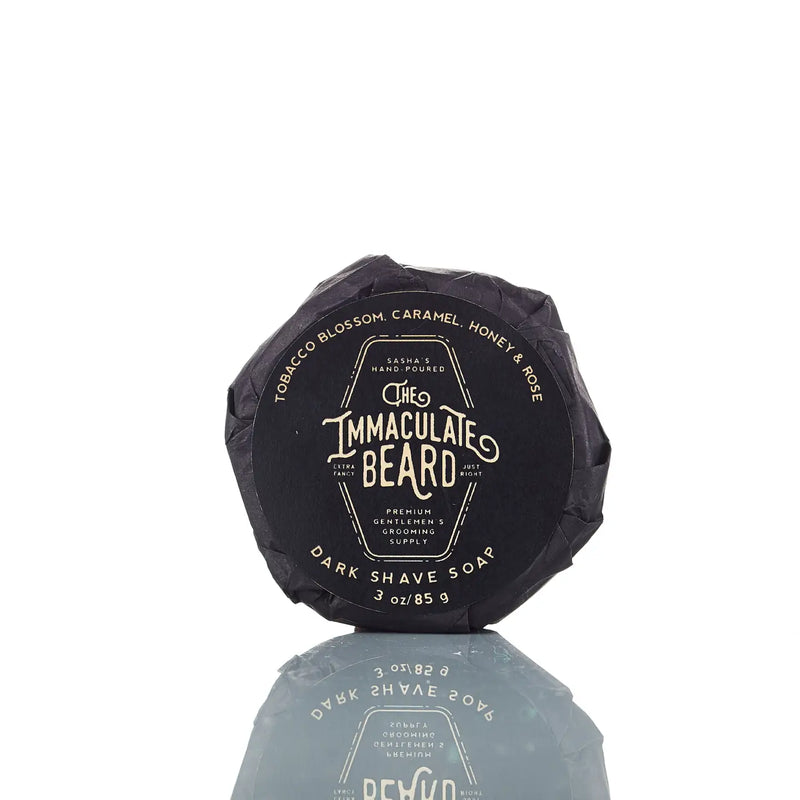 The Immaculate Beard DARK Shave Soap Puck, ideal for sensitive skin, comes in a black paper wrap with gold and white text highlighting its scents: tobacco blossom, caramel, honey, and rose.
