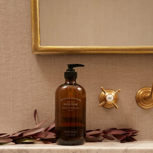 A brown glass bottle of Archipelago Botanico de Havana Hand Wash by Archipelago Botanicals rests on a bathroom counter near a gold faucet handle, burgundy leaves, and a gold-framed mirror.