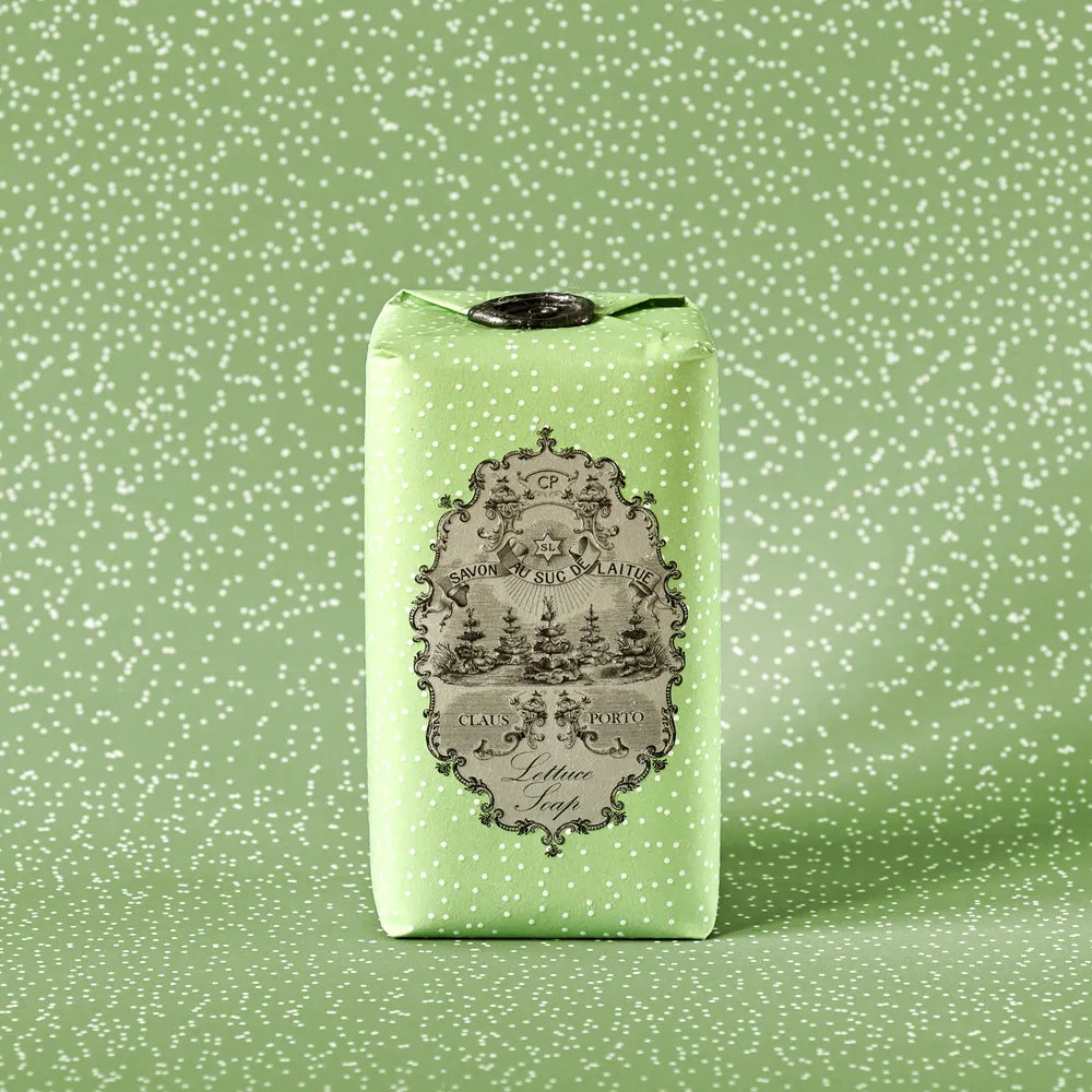A bar of Claus Porto 1887 Classico Spring Lettuce Soap, wrapped in light green paper with white polka dots, features an ornate black-and-white vintage label showcasing pistachio nut seed oil and a scenic illustration with decorative text.