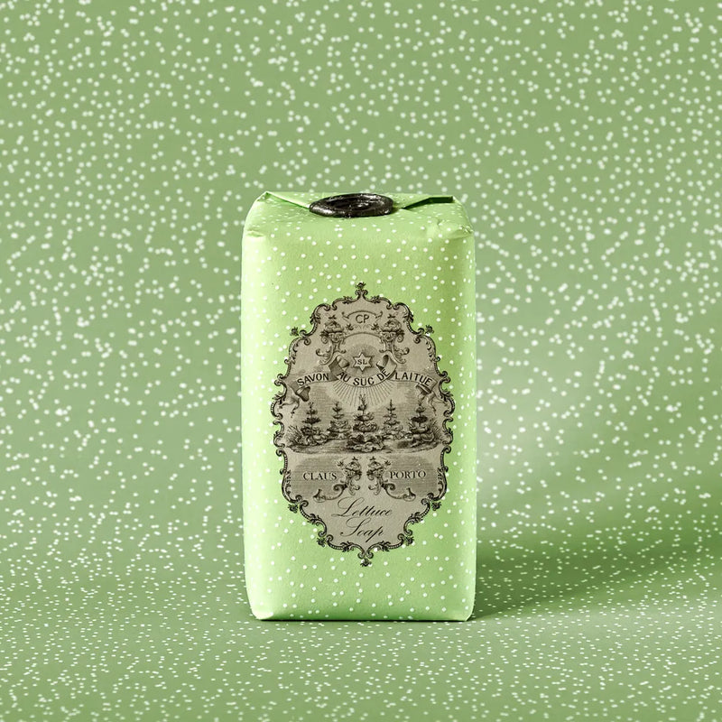 A bar of Claus Porto 1887 Classico Spring Lettuce Soap, wrapped in light green paper with white polka dots, features an ornate black-and-white vintage label showcasing pistachio nut seed oil and a scenic illustration with decorative text.