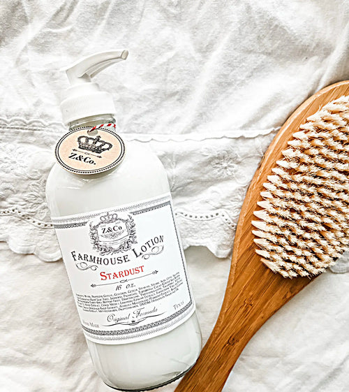 A bottle of Z&Co. Stardust Farmhouse Lotion featuring natural ingredients sits on white lace fabric beside a wooden brush with soft bristles, creating a rustic and elegant skincare vibe.