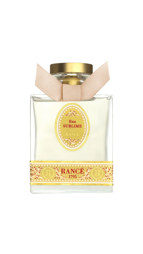 Rancé Sublime Eau de Parfum by Rancé comes in a square glass bottle with a gold cap and beige ribbon, adorned with yellow ornate details that echo the refined elegance of its floral fragrance.