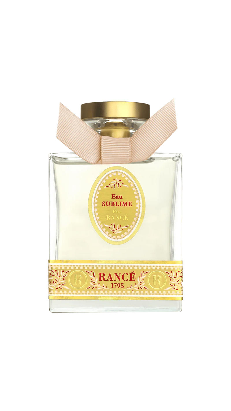 Rancé Sublime Eau de Parfum by Rancé comes in a square glass bottle with a gold cap and beige ribbon, adorned with yellow ornate details that echo the refined elegance of its floral fragrance.
