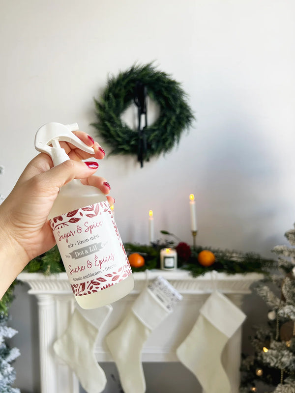 A hand with red-painted nails holds a Dot & Lil Sugar & Spices Linen + Air Mist in front of a festive mantel with stockings, candles, greenery, and a wreath—ready to fill your home with the scent of spiced cookies.