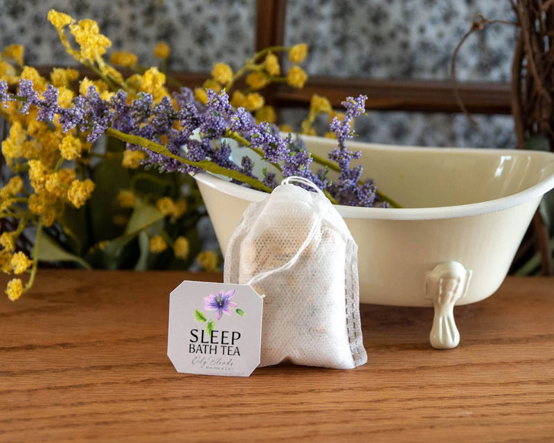 A small white clawfoot bathtub holds a bouquet of purple and yellow flowers. In front, a labeled "Hampton Court Bath Tea Single Bag - Sleep," infused with essential oils, rests on a wooden surface. The floral background enhances the cozy ambiance, inviting relaxation.