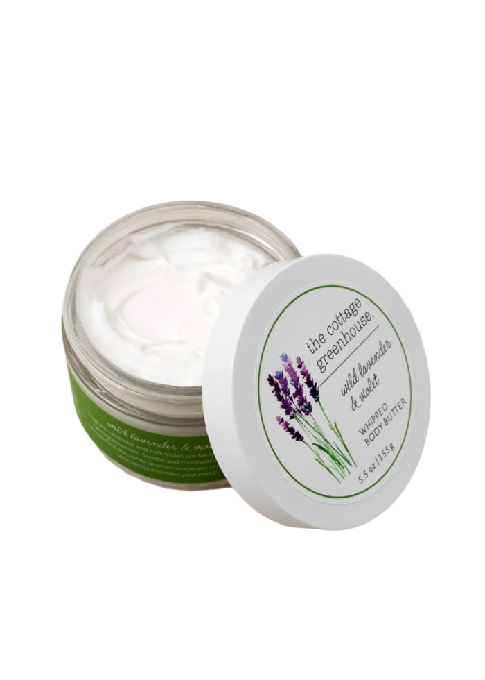 An open container of The Cottage Greenhouse Wild Lavender & Violet Body Butter by Margot Elena comes with a white lid featuring lavender illustrations. Made with natural ingredients, this creamy body butter offers a nourishing and moisturizing experience in its green and white packaging.