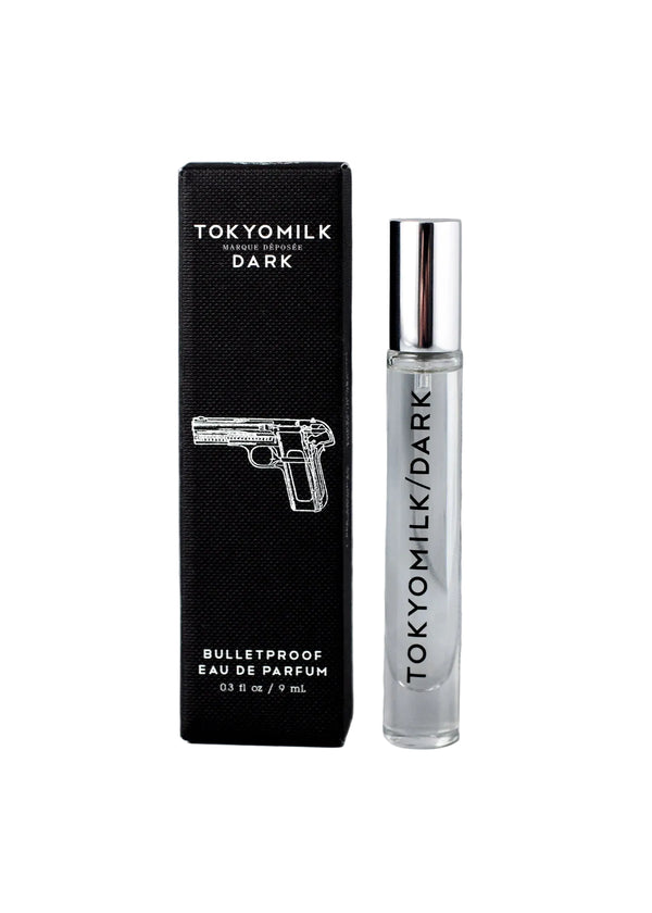 A clear glass bottle of Margot Elena’s TokyoMilk Dark Bulletproof Travel Eau de Parfum sits beside a sleek black box with a gun illustration and bold text, emphasizing this distinctive unisex fragrance.