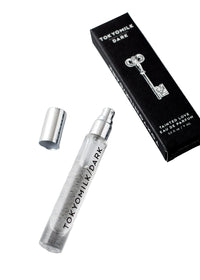 A clear glass spray vial of Margot Elena’s TokyoMilk Dark Tainted Love Travel Eau De Parfum with silver cap rests next to a black box displaying a white key and label, highlighting dark vanilla bean notes.