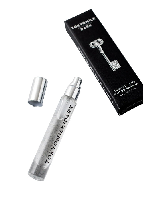 A clear glass spray vial of Margot Elena’s TokyoMilk Dark Tainted Love Travel Eau De Parfum with silver cap rests next to a black box displaying a white key and label, highlighting dark vanilla bean notes.