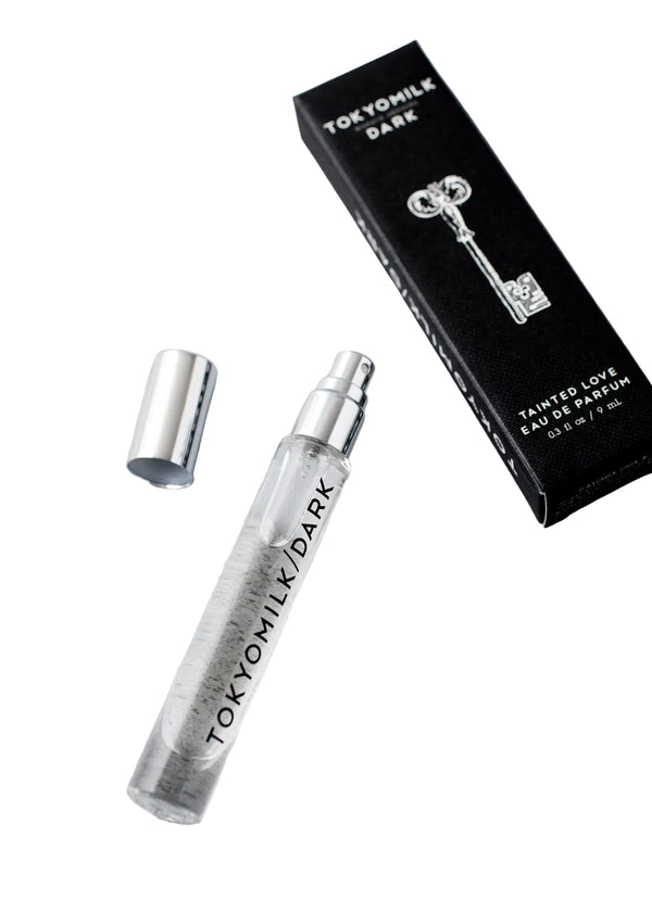 A clear glass spray vial of Margot Elena’s TokyoMilk Dark Tainted Love Travel Eau De Parfum with silver cap rests next to a black box displaying a white key and label, highlighting dark vanilla bean notes.
