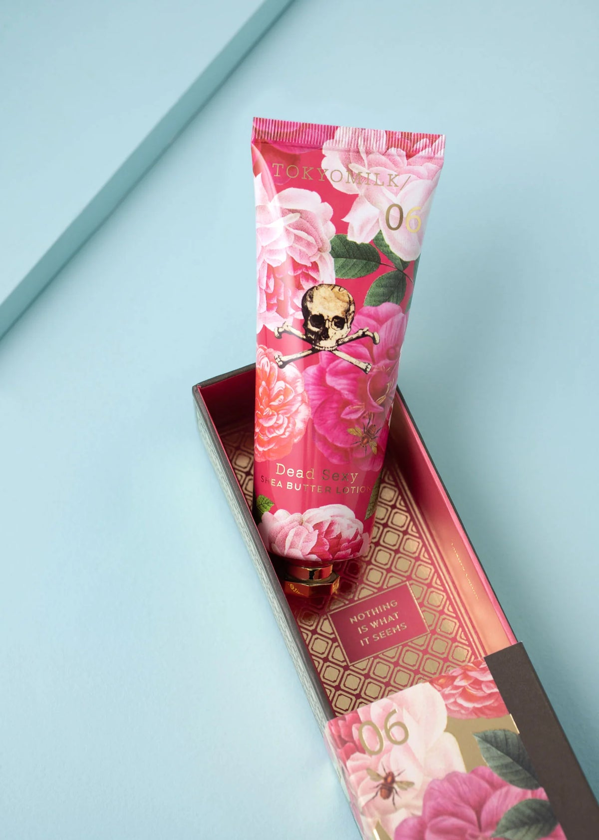 A floral tube labeled "TokyoMilk Dead Sexy Large Handcreme" by Margot Elena, featuring a skull and rose design, rests in an open decorative box on a light blue surface, offering intense hydration for hands.