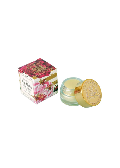 A small glass jar of Margot Elena's TokyoMilk Honeyed Apricot Lip Balm with a gold lid sits beside its decorative floral box, both displayed on a white background.