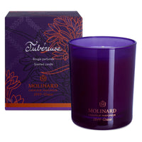 The Molinard Tuberose Candle sits next to its deep purple box adorned with floral designs, orange accents, and gold branding from Molinard, adding a touch of luxury to your home décor.
