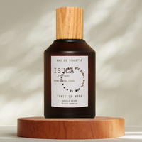 A dark brown glass perfume bottle with a wooden cap rests on a light round platform. The label reads "ISULA parfums Vanilla Noir eaux de toilette," showcasing this vanilla fragrance by ISULA parfums as sunlight casts gentle shadows behind it.