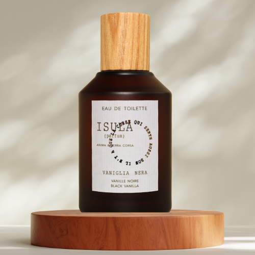 A dark brown glass perfume bottle with a wooden cap rests on a light round platform. The label reads "ISULA parfums Vanilla Noir eaux de toilette," showcasing this vanilla fragrance by ISULA parfums as sunlight casts gentle shadows behind it.