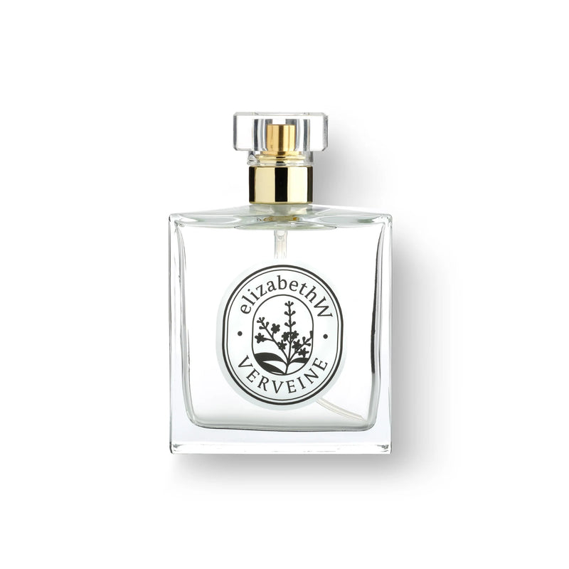 A square glass perfume bottle with a gold cap, featuring a white label with black text "elizabeth W Atelier Verveine Eau de Parfum" and a decorative emblem. The background is.