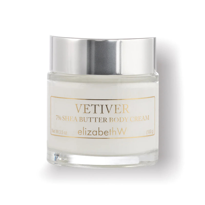 A jar of elizabeth W Signature Vetiver Body Cream elegantly showcases the luxurious shea butter inside. The clear container reveals the smooth, white cream, complemented by a silver lid and a chic white label featuring gold and gray text.