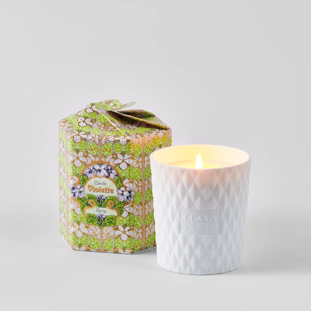 The Claus Porto 1887 Ltd Edition Violette Spring Flowers Candle, in textured white Portuguese porcelain, sits by its floral box and fills the light gray space with a delicate floral-oriental scent.
