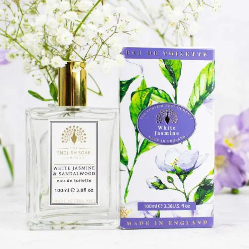 A clear perfume bottle labeled "The English Soap Co. White Jasmine Eau de Toilette" stands next to its floral-patterned box with gold and purple accents. This cruelty-free scent, made in England, is surrounded by white flowers.