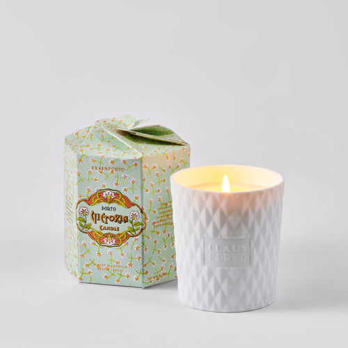 A Claus Porto 1887 Ltd. Edition Victoria White Magnolia Candle glows in a textured biscuit porcelain holder embossed with “Claus Porto,” beside its pastel green floral “Porto” box.