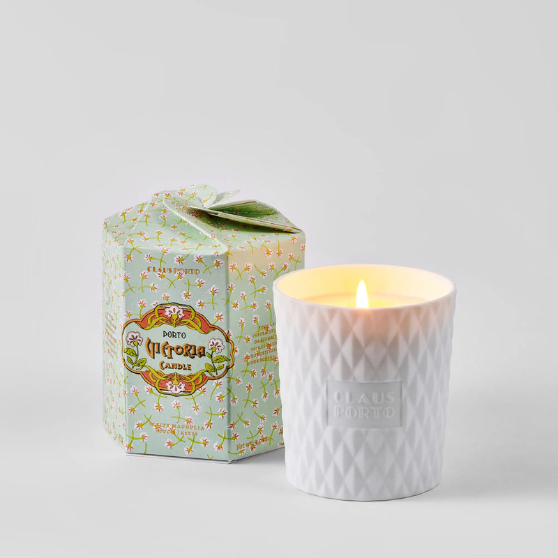A Claus Porto 1887 Ltd. Edition Victoria White Magnolia Candle glows in a textured biscuit porcelain holder embossed with “Claus Porto,” beside its pastel green floral “Porto” box.