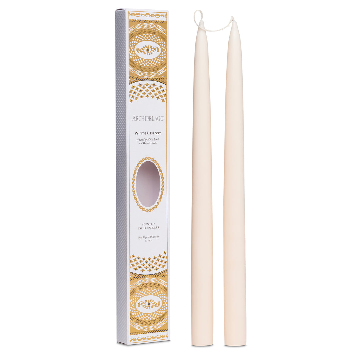 Two tall Winter Frost taper candles by Archipelago Botanicals stand next to a decorative white and gold box labeled "Archipelago Winter Frost Tapered Candle Set," featuring an oval window revealing the candles inside.