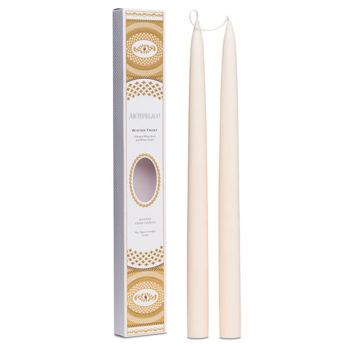 Two tall Winter Frost taper candles by Archipelago Botanicals stand next to a decorative white and gold box labeled "Archipelago Winter Frost Tapered Candle Set," featuring an oval window revealing the candles inside.