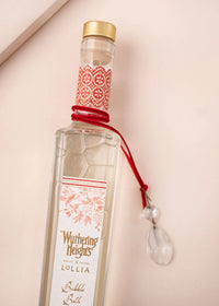 A tall, clear bottle of Lollia "WUTHERING HEIGHTS" Kiss Me Again Bubble Bath by Margot Elena, with gold and red accents, a red wax seal, and a glass pendant on the neck, exudes luxury against a light background.