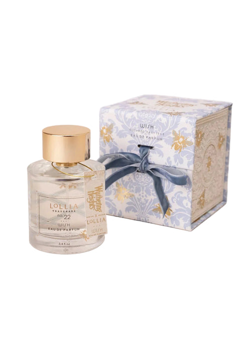A clear glass Lollia ‘WUTHERING HEIGHTS’ Wish Eau de Parfum bottle by Margot Elena with a gold cap sits beside an ornate blue and white box with a blue ribbon, evoking literary romance against a soft pink background.