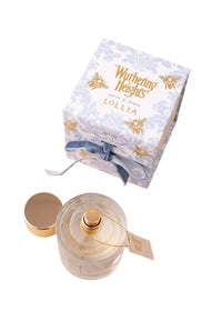 A clear glass Margot Elena Lollia 'WUTHERING HEIGHTS' Wish Eau de Parfum bottle with a gold cap rests open beside an ornate blue and white box accented with gold and a blue ribbon, on a pale pink surface.